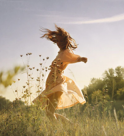 A person with long hair jumping joyfully in a field at sunset, wearing a flowing dress, with wildflowers in the foreground and golden sunlight creating a dreamy atmosphere