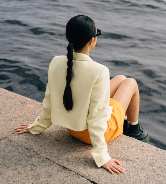 A person sitting on a concrete surface by the water, wearing a cream colored long-sleeve top and orange shorts, with long dark hair in a braid, viewed from behind
